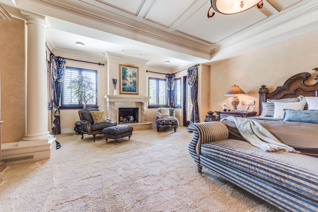 Spacious bedroom designed by a Vancouver general contractor, featuring a fireplace, ornate furniture, large windows, and luxurious decor in soft beige and blue tones.
