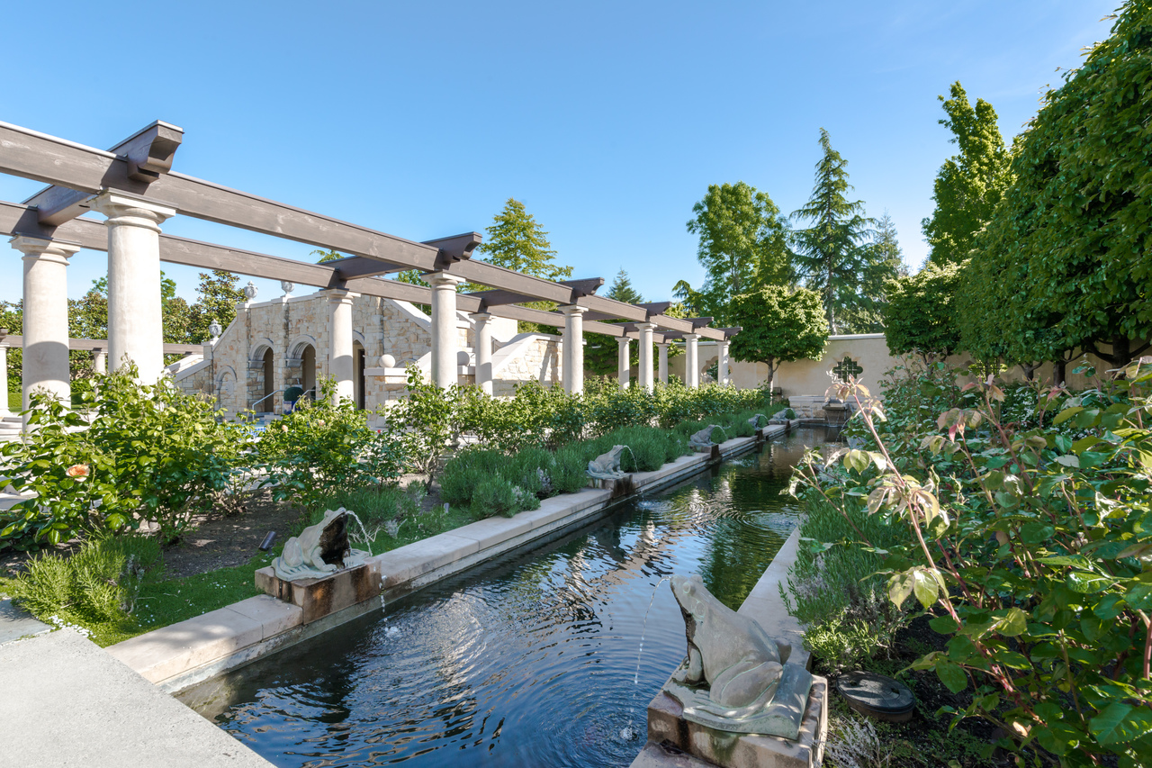 Rectangular fountain with frog statues, surrounded by a lush garden and stone columns under a clear blue sky—an elegant construction inspired by classic Vancouver gardens in British Columbia.