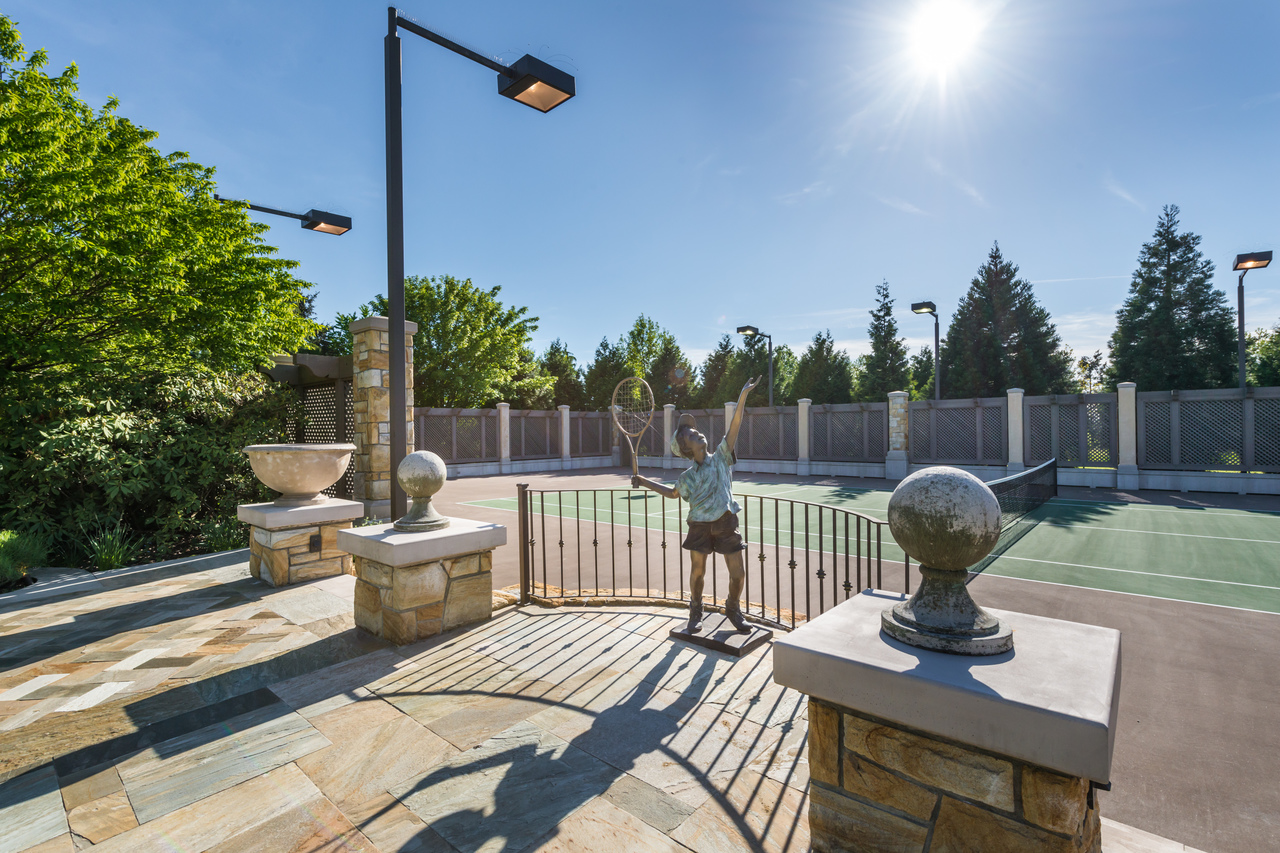 Bronze tennis player statue by a court gate in Vancouver, sun shining, with trees and lamps in the background—crafted with skill often seen from a top BC general contractor.