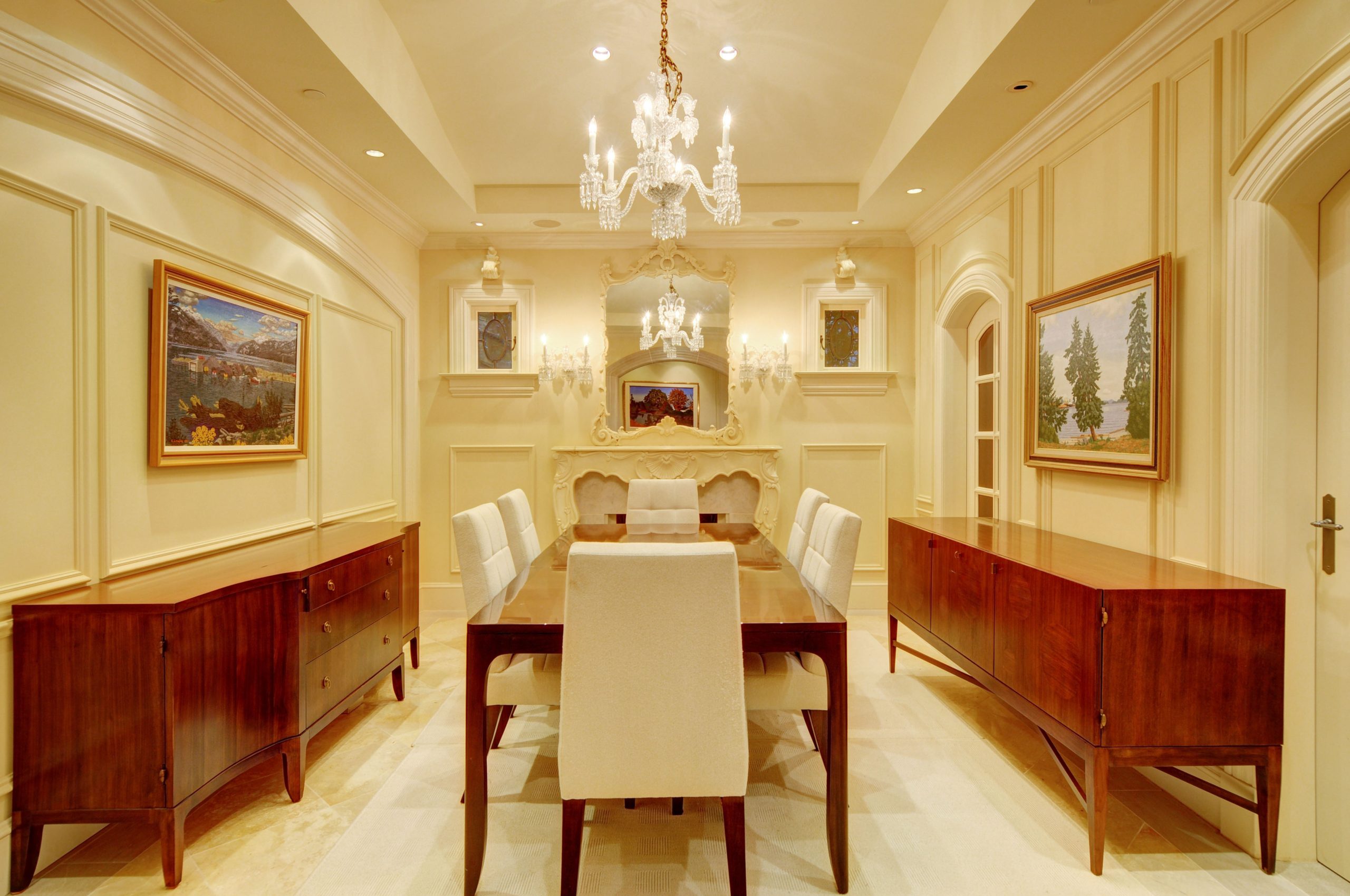 Dining room in Vancouver with a wooden table, white chairs, chandeliers, and framed art on beige walls.