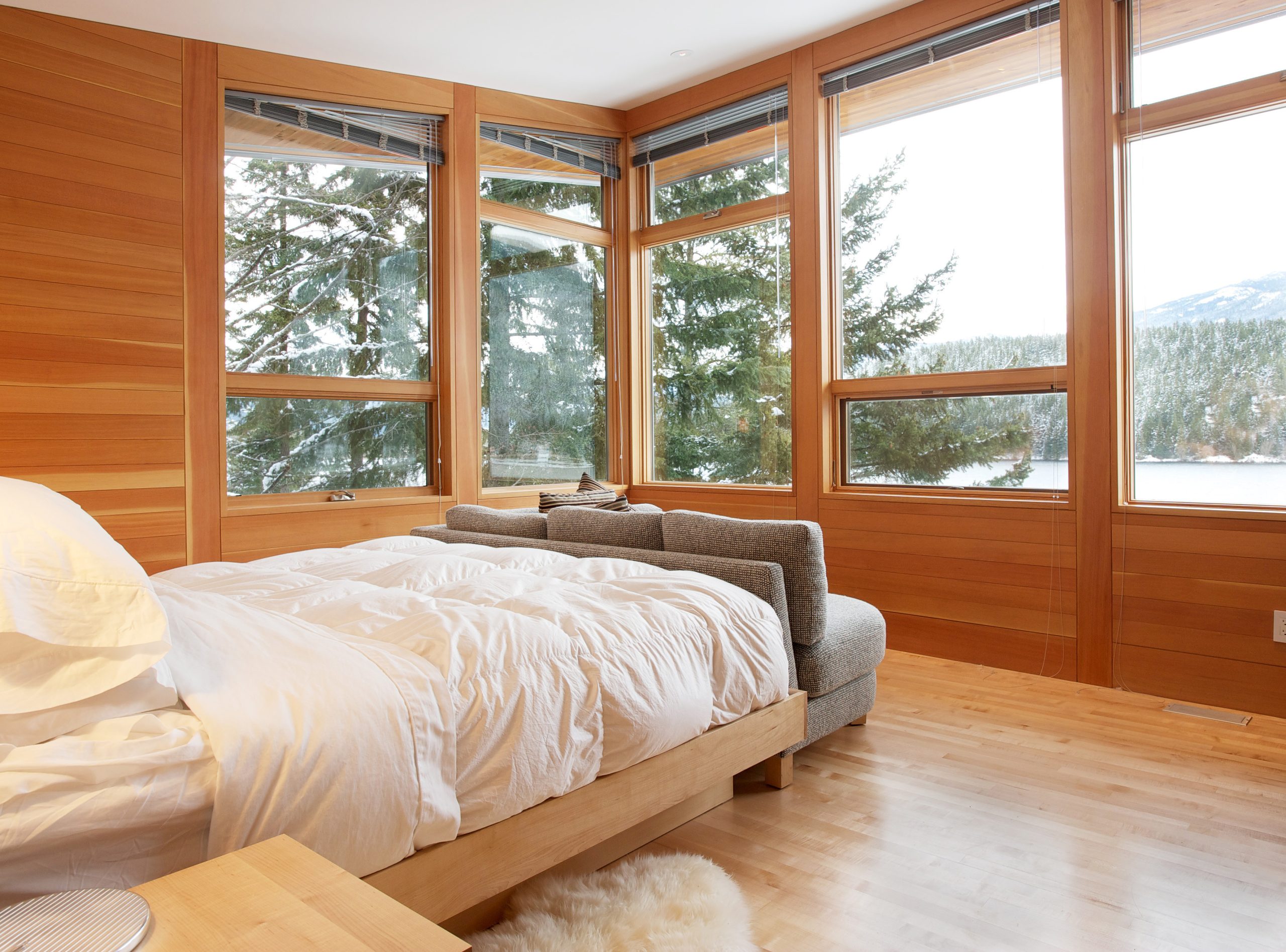 Bedroom with wooden walls, large windows, white bedding, and a snowy forest view outside—crafted by a leading Vancouver general contractor for timeless comfort.