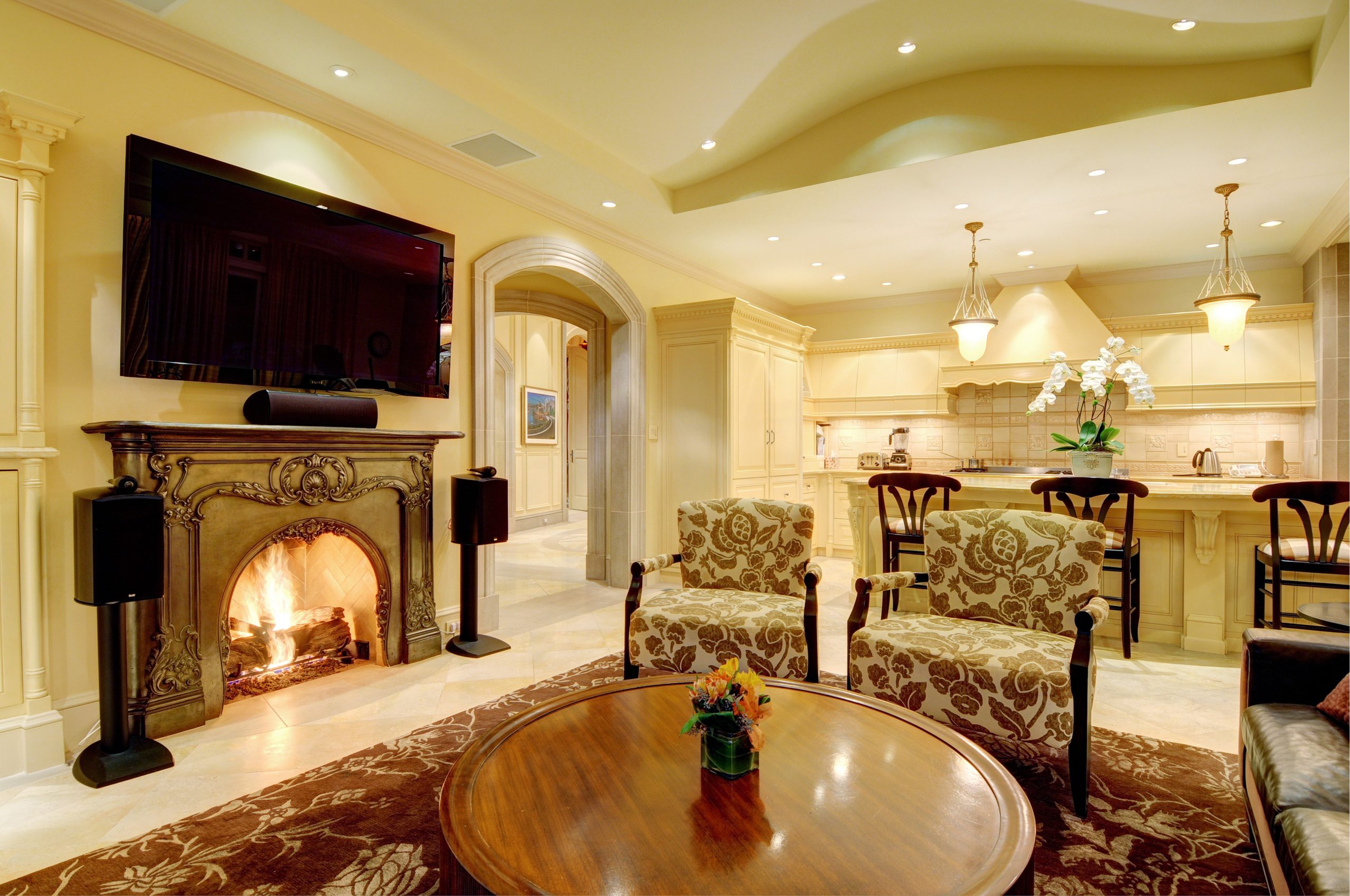 Living room with quality architecture, featuring a fireplace, patterned chairs, a TV above the mantel, and an open kitchen in view—an inviting space inspired by British Columbia’s charm.