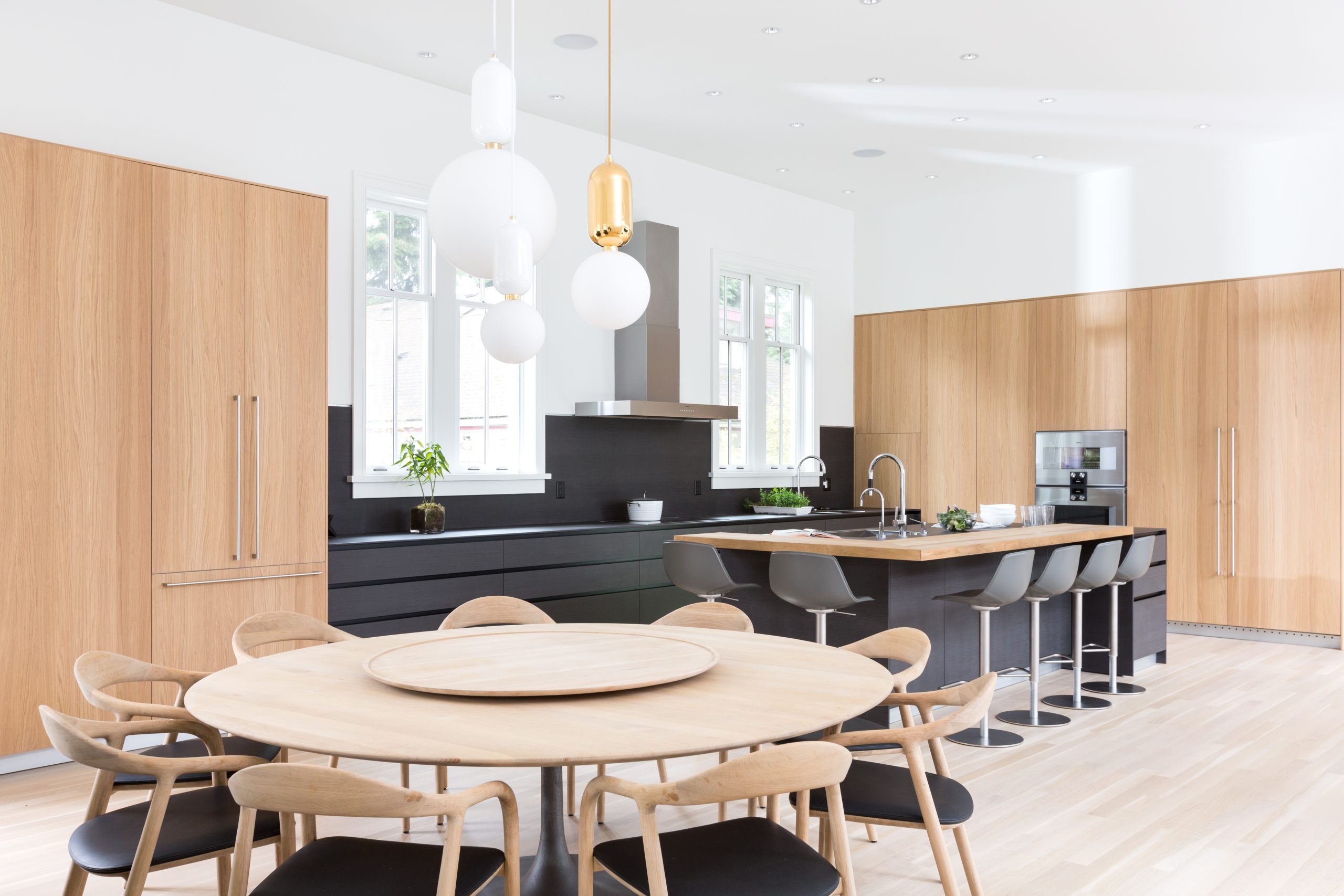 Sleek kitchen with wooden cabinets, black island, round dining table, and hanging pendant lights—showcasing custom homes architecture inspired by British Columbia.