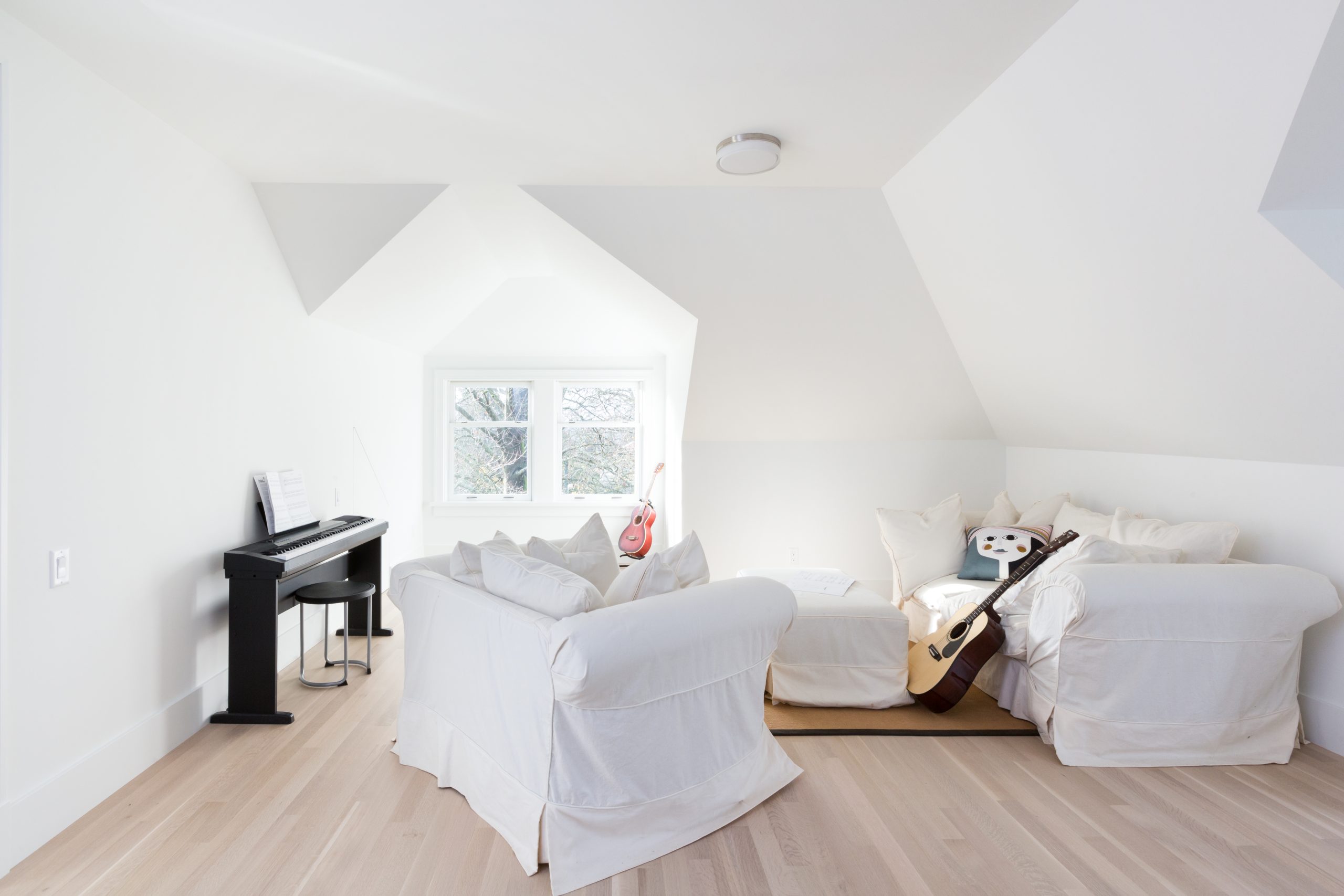 Bright attic room in Vancouver featuring white couches, a keyboard, guitar, and a large window letting in natural light—perfect for custom homes that blend comfort and quality.