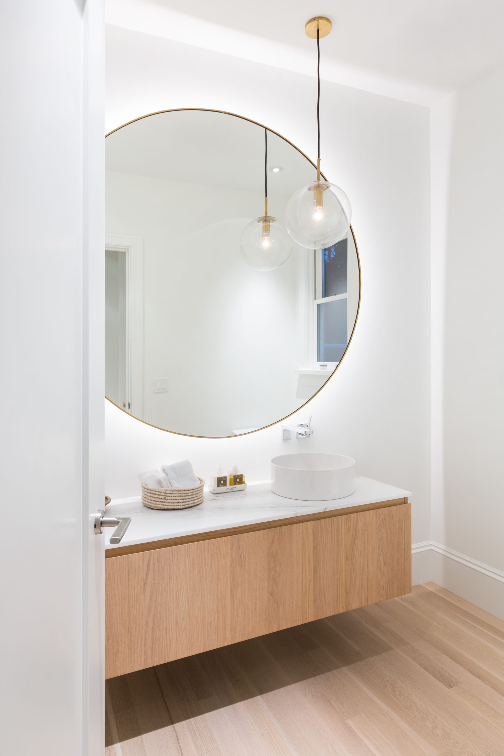 Minimalist bathroom in Vancouver, BC, featuring a round mirror, wooden vanity, vessel sink, and two globe pendant lights.