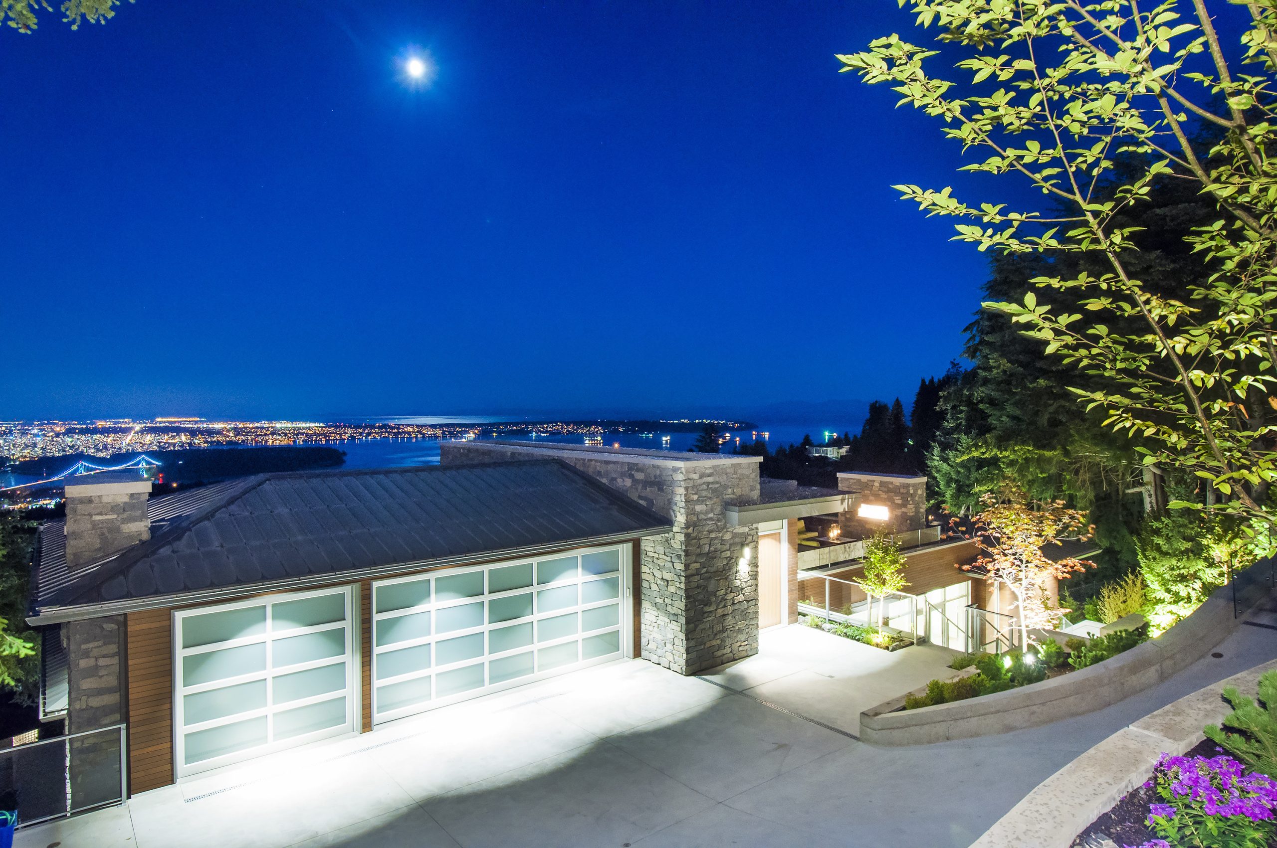 House with striking architecture and large garage at night, overlooking Vancouver’s city lights and water under a bright moonlit sky.