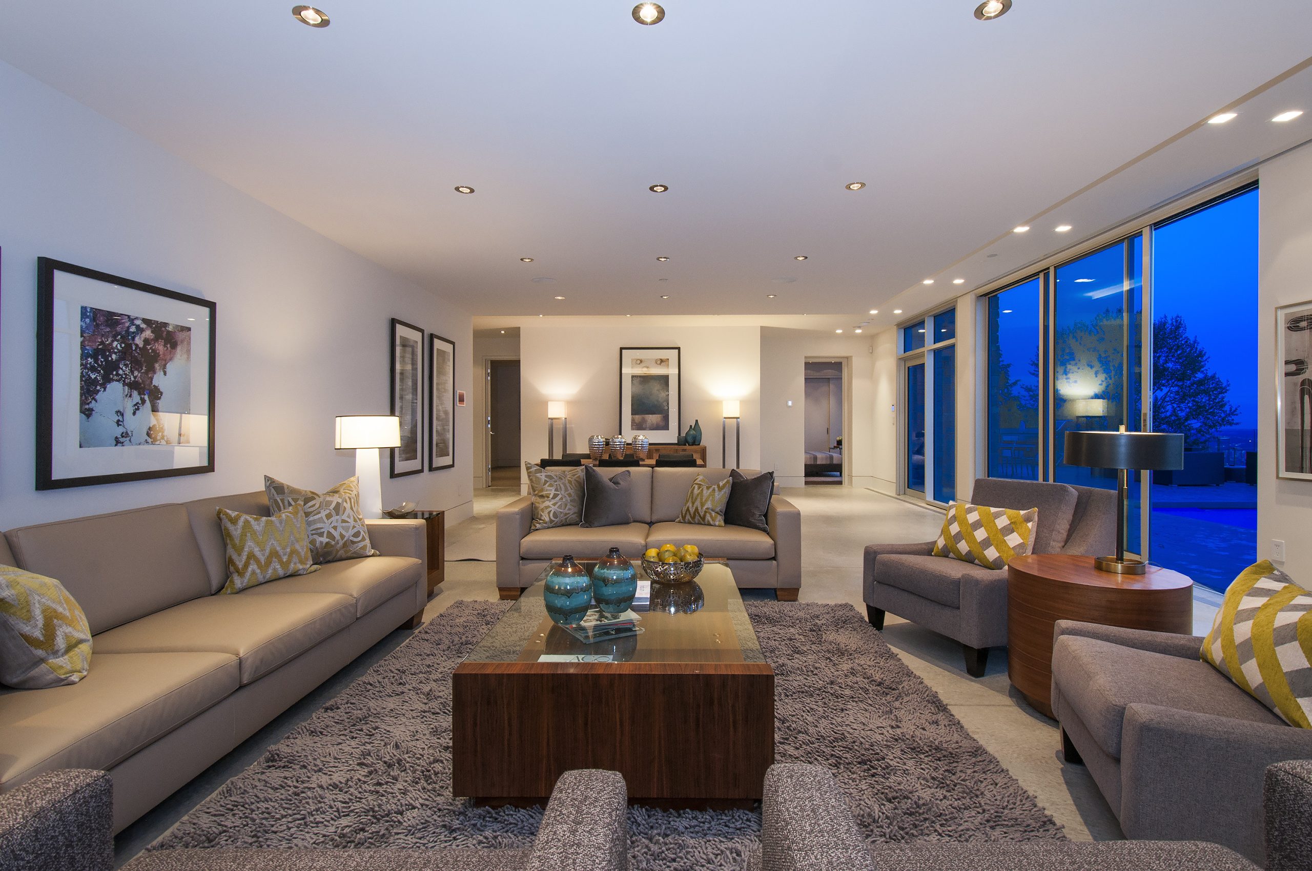 Living room in a custom home, featuring neutral sofas, patterned pillows, art on the walls, large windows, and a blue evening sky—showcasing refined architecture and quality throughout.
