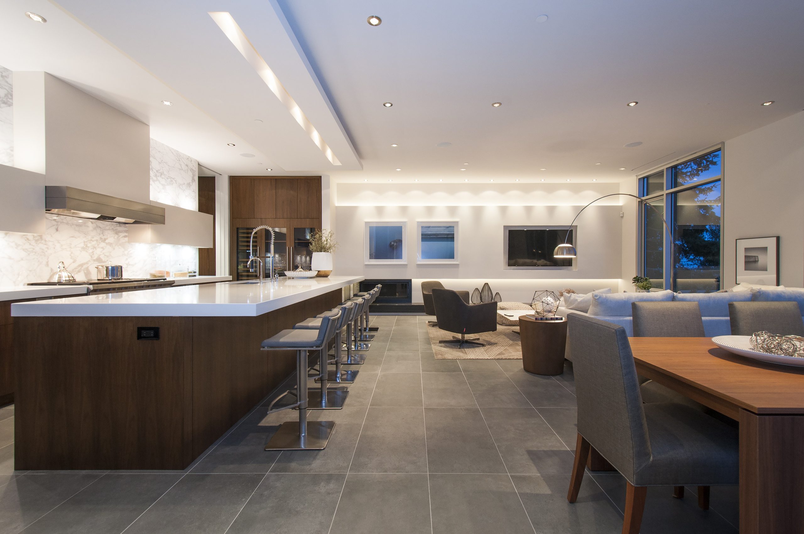Open-concept kitchen and living area with island seating, grey floors, and large windows at dusk—crafted by a reputable company specializing in custom homes across British Columbia.