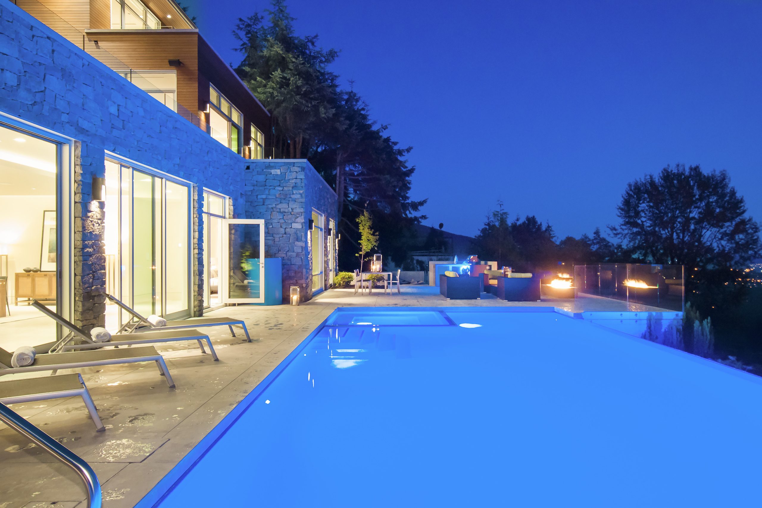 House with large windows and a glowing pool at dusk, surrounded by lounge chairs and outdoor lights—a stunning example of custom homes in British Columbia.