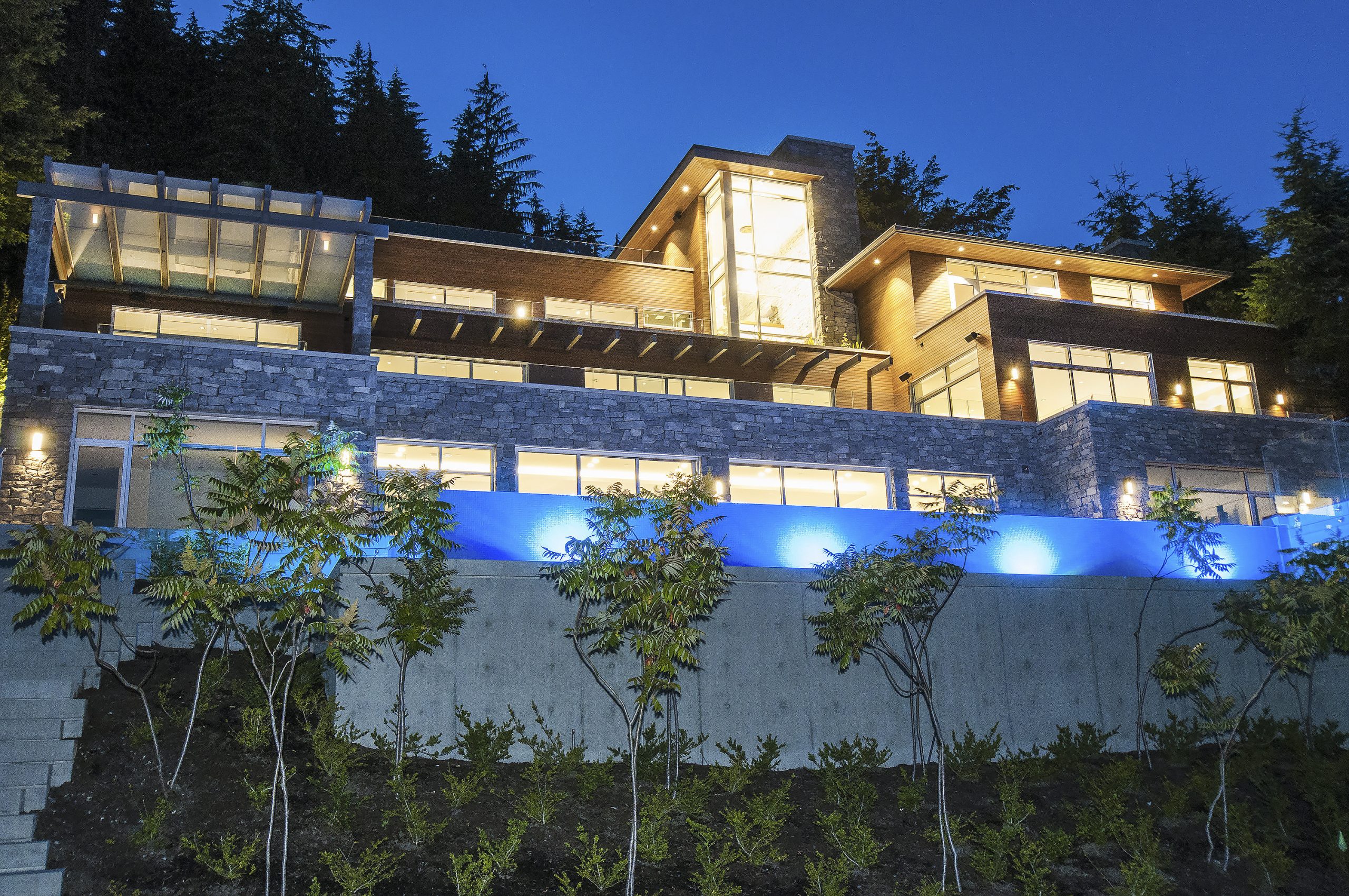 Modern multi-story house with large windows, stone facade, and blue lighting, surrounded by trees at dusk—an example of quality custom homes built by a reputable company based in Vancouver