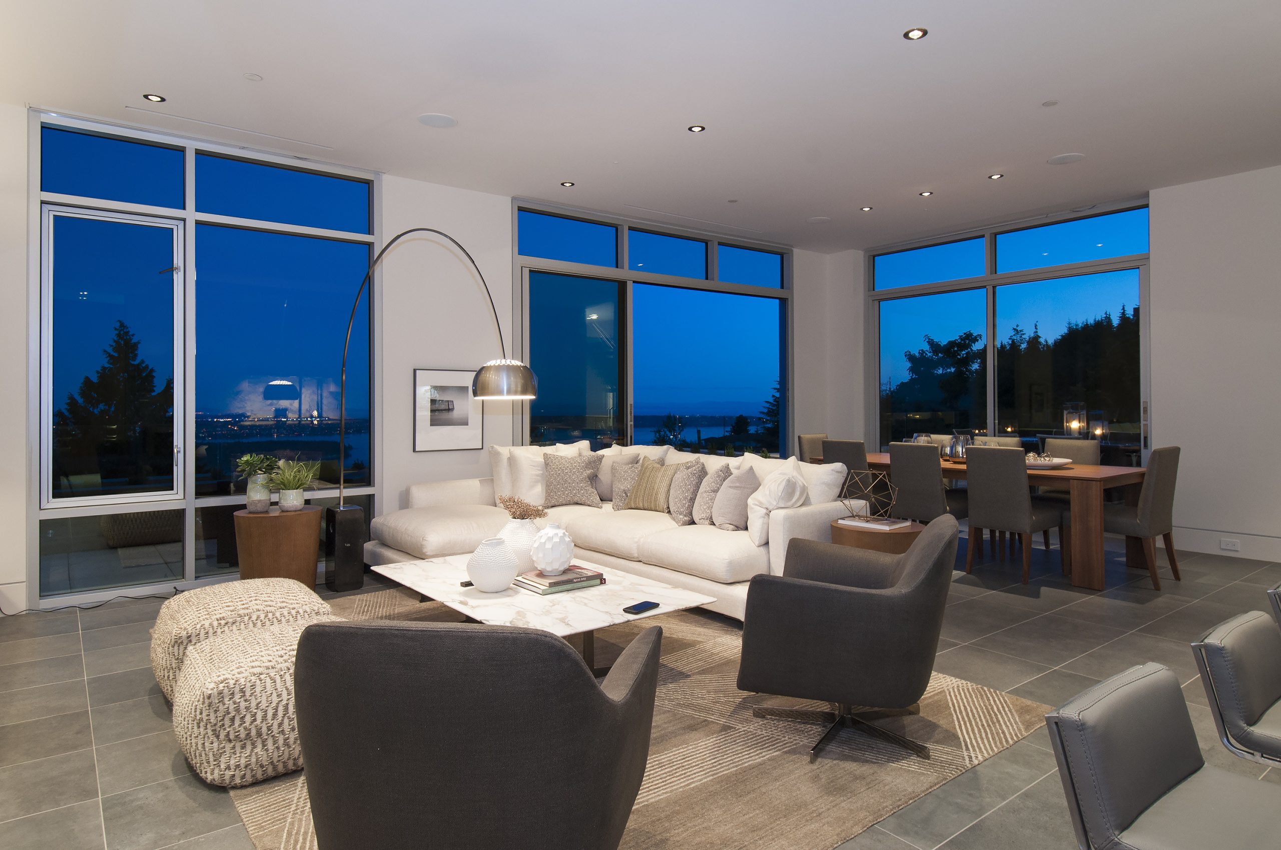 Living and dining room with large windows, quality gray furniture, and a stunning view of Vancouver at dusk.