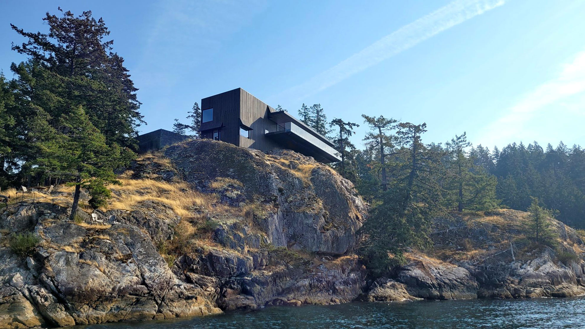 Black house sits atop a rocky, tree-covered cliff overlooking the water under a clear blue sky, showcasing sleek construction inspired by British Columbia’s stunning natural landscapes.