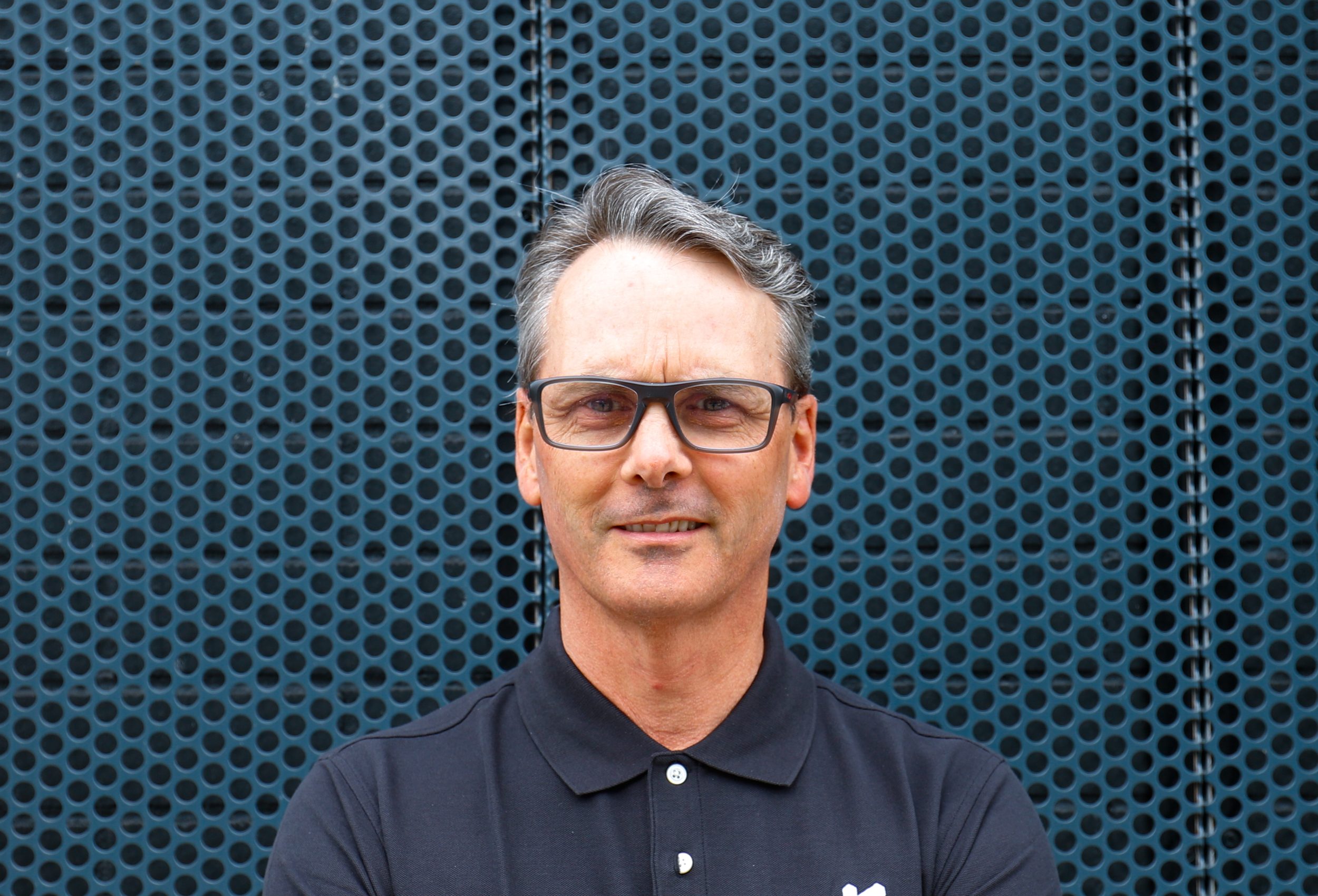 Man with glasses and gray hair wearing a black polo shirt stands in front of a perforated metal wall, embodying the professionalism of a BC construction expert.