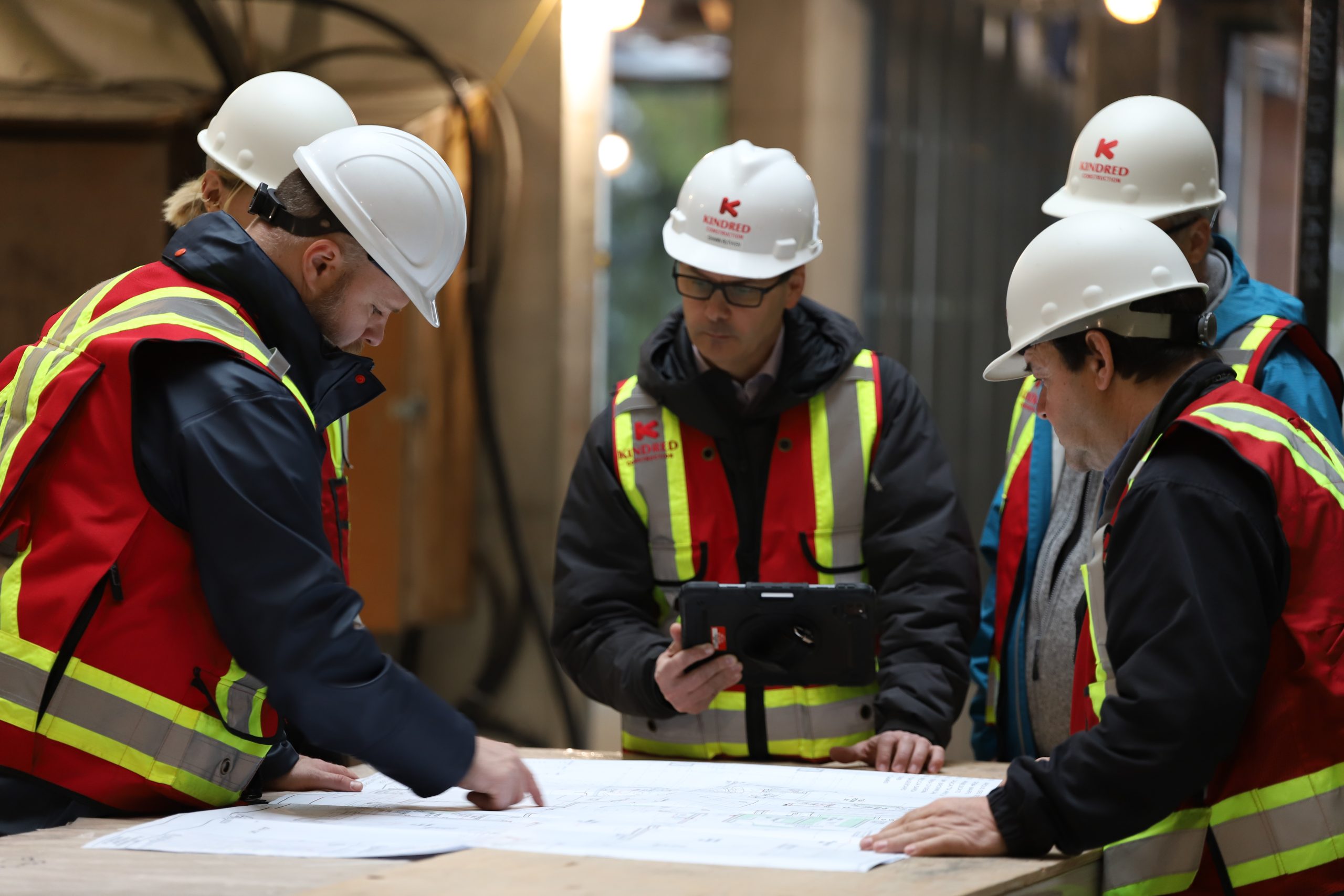 Five construction workers in safety gear review blueprints at a table on a worksite indoors.